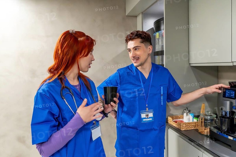 Healthcare professionals wearing blue scrubs on a break, preparing coffee while talking and smiling, showing teamwork and a positive work environment in a hospital setting