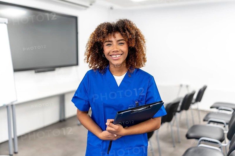 Young female medical student dressed in blue scrubs holding a clipboard while standing confidently and smiling in an empty classroom, ready for a lecture or training session