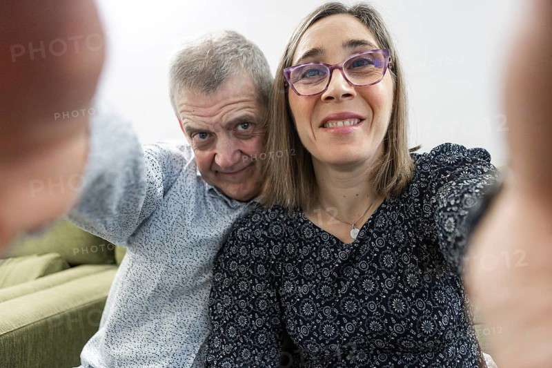 A mature couple with intellectual disabilities is joyfully taking a selfie while sitting on a cozy couch at supervised home. The relaxed setting, inviting atmosphere