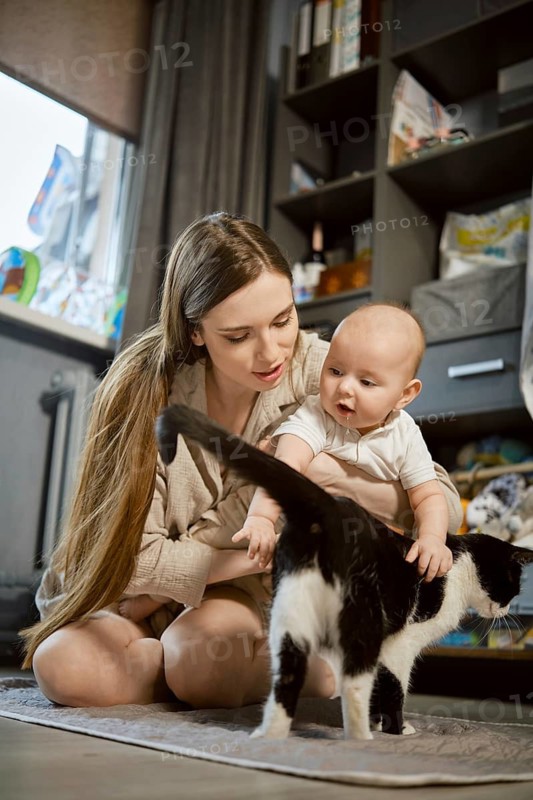 A mother kneels on the floor in a living space while her baby sits next to her. The baby looks at a black and white cat, reaching out to touch its tail. Both are engaged in this moment