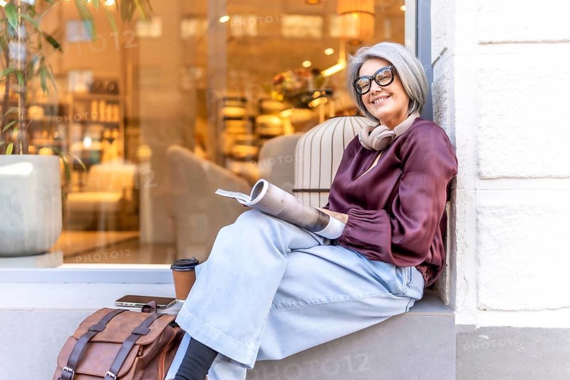 Smiling mature woman with grey hair and eyeglasses sitting on a city ledge and reading a magazine, enjoying a relaxed moment with coffee and headphones nearby