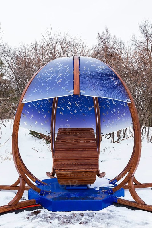 Interactive cocoon rocking chair made of curved wood and backlit blue tinted plexiglass panels canopy decorated with flock of birds in flight theme motifs created by Olivier Landreville and installed in winter like snow in early spring, Horizon Art Installation, Ile des Moulins, Old Terrebonne, Quebec, Canada