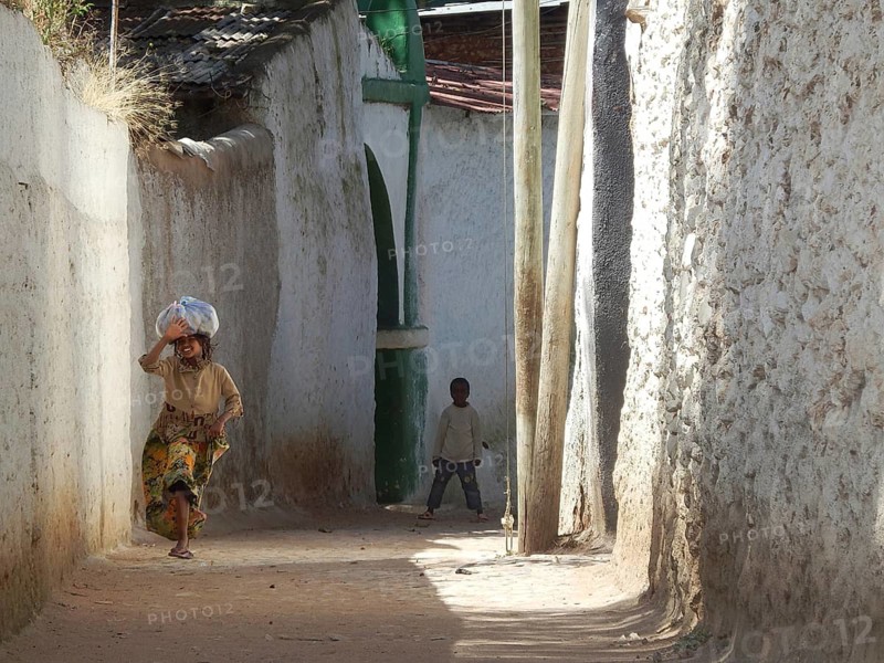 A woman carries a package on her head in a narrow alley and waves while a child watches, Harar, Ethiopia