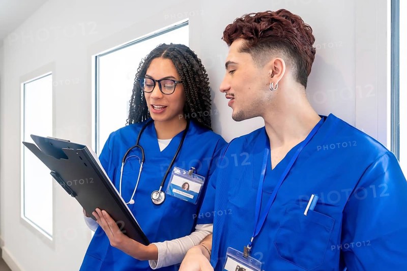 Young medical professionals wearing blue scrubs and stethoscopes, discussing information on a clipboard while standing in a brightly lit clinic hallway, collaborating on patient care