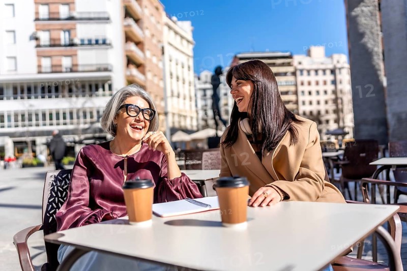 Two women laughing and talking over coffee at an outdoor cafe table in a sunny urban setting, sharing ideas and enjoying friendship during a relaxed downtown meeting