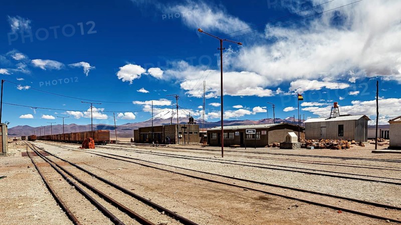 Lonely train stop with scattered buildings and clouds in the sky in front of a mountain range, the town of Ollagüe in the Chilean Atacama Desert with the Ollagüe volcano of the same name in the background