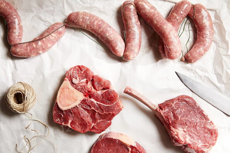 A variety of fresh meat cuts and linked sausages are elegantly arranged on butcher paper, alongside a sharp knife and twine, evoking the art of traditional butchery