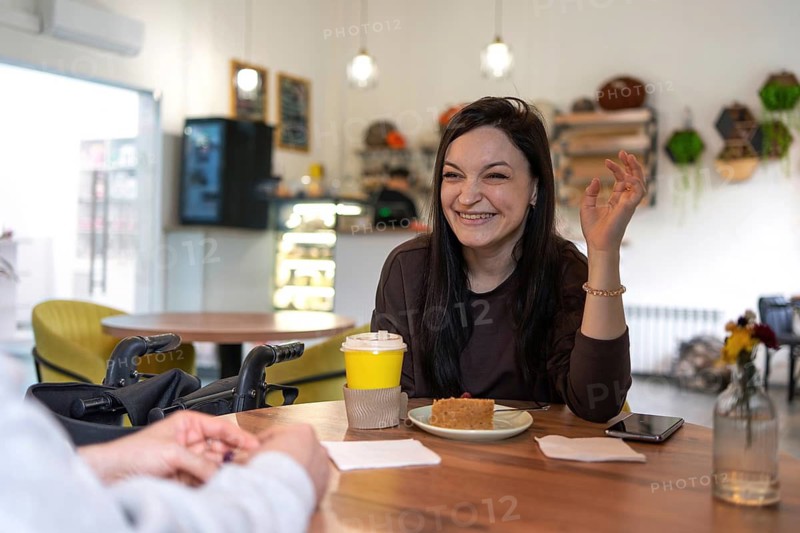 A woman with cerebral palsy is smiling while enjoying coffee and cake in a cozy cafe setting. She is engaged in conversation, creating a warm and inviting atmosphere