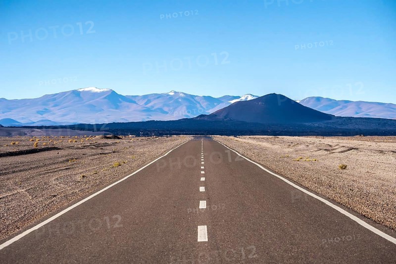 A straight road cuts through the arid desert landscape of La Puna in Argentina, with snow-capped mountains and scattered vegetation under a clear sky