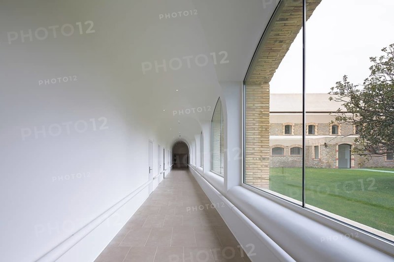 A sleek, white corridor of hospital with arched windows offering a view of a historic brick hospital and lush green lawn outside. The minimalist design adds elegance and tranquility