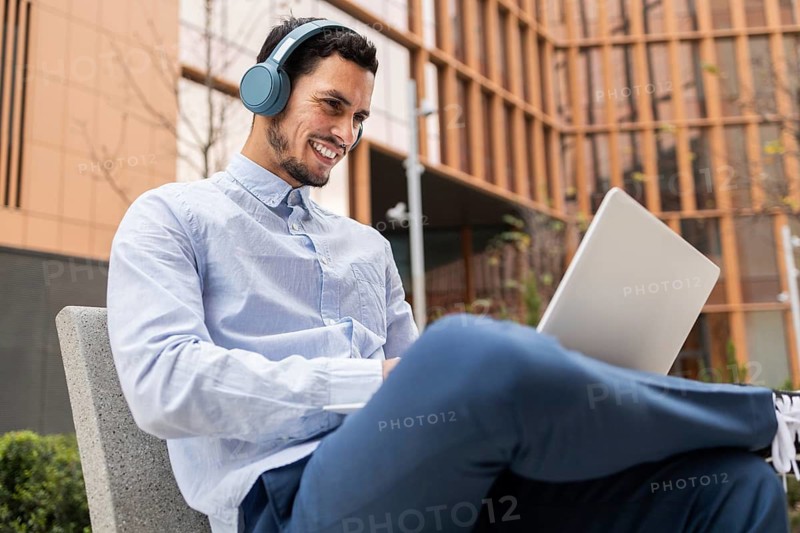 A young man in business casual attire sits outdoors with a laptop and headphones, smiling while working. The modern urban background highlights productivity, remote work, and digital connectivity