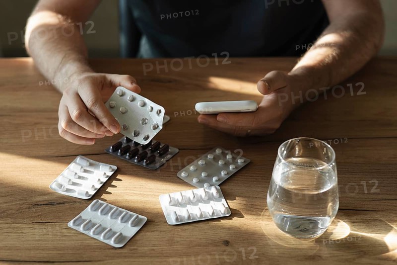 A man holding a blister pack of pills while using a smartphone, with several packs and a glass of water on a wooden table, highlighting medication management at home