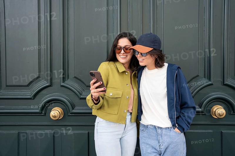 A cheerful lesbian couple takes a selfie together, standing close with smiles, dressed casually in stylish jackets and sunglasses. They radiate happiness and love