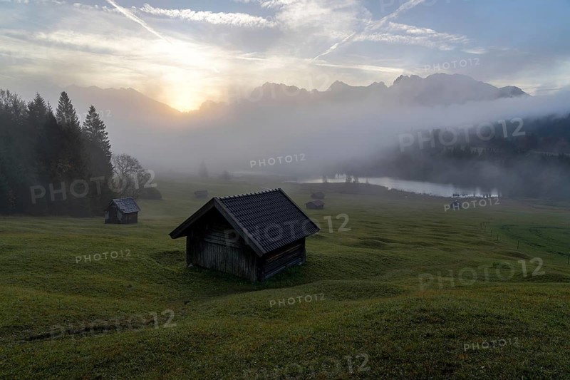 A serene view of Geroldsee in the Bavarian Alps at sunrise, featuring misty fields, rustic wooden huts, and the majestic silhouette of mountains under a glowing sky
