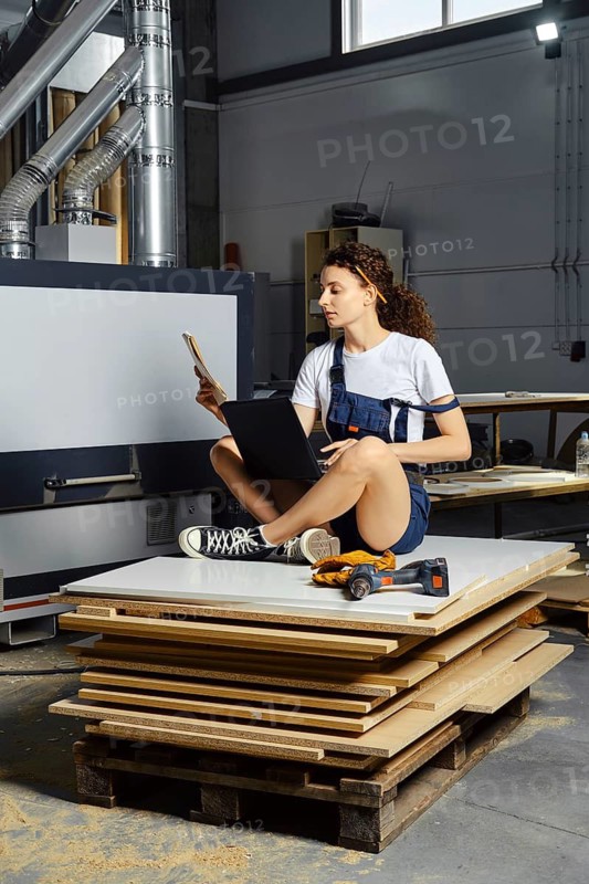 Young woman in uniform sits on a stack of chipboard slabs with laptop and paper blueprint in hand