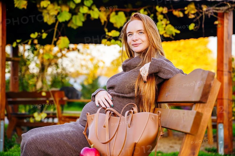 A beautiful pregnant woman with long red hair sits leisurely on a wooden bench outdoors, enjoying the warmth and serenity of a sunny day surrounded by lush foliage