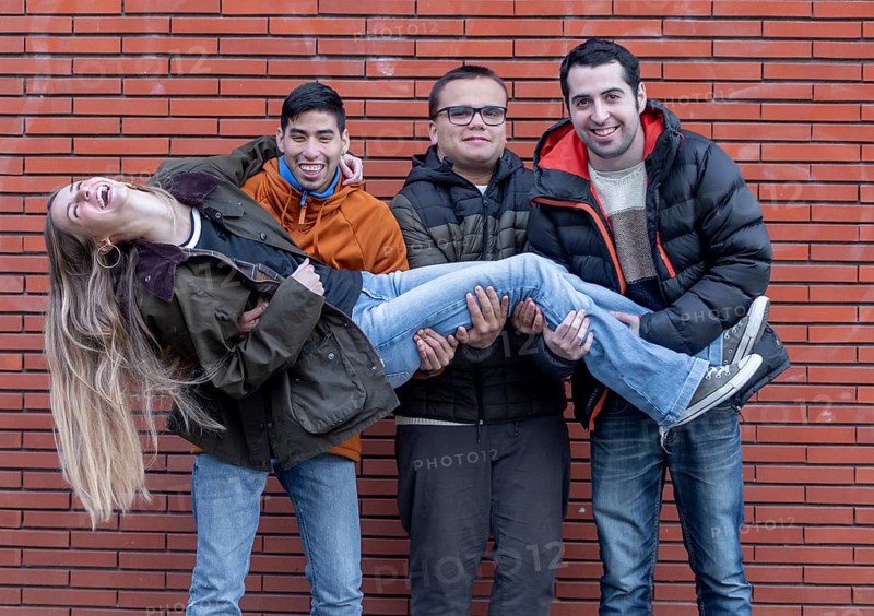 A joyful group at a mental health leisure center shares a playful moment as three young men with intellectual disabilities lift their smiling caregiver