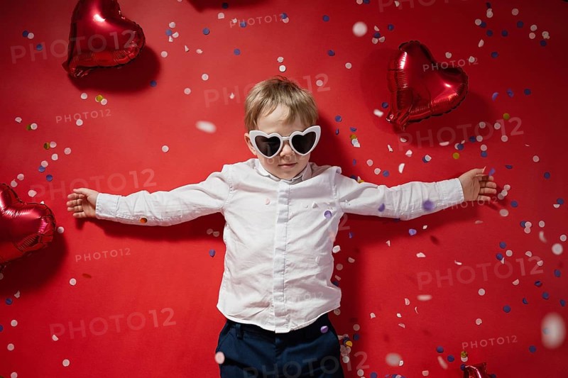 A young boy with heart-shaped sunglasses stands with outstretched arms surrounded by red heart-shaped balloons and colorful confetti against a vibrant red background