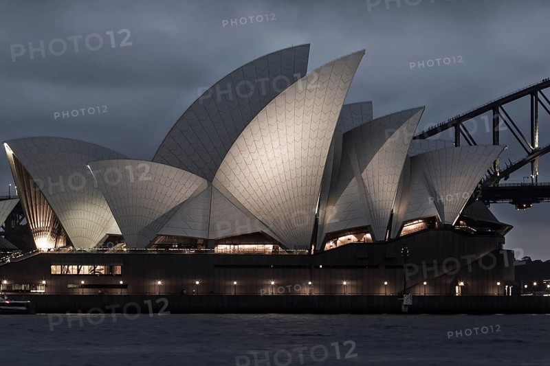 A stunning view of the Sydney Opera House illuminated against the night sky. The architectural masterpiece shines with lights, creating a captivating glow amid the cloudy evening