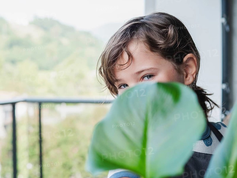 View of a young girl playfully hiding behind a large green leaf on a balcony, looking at camera, with a blurred nature background