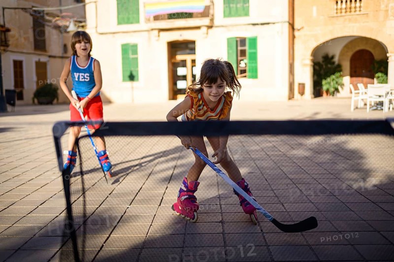 Two children playing roller hockey on roller blades in a sunny urban plaza, displaying joy and teamwork in an outdoor recreational activity. The focus is on fun and active play