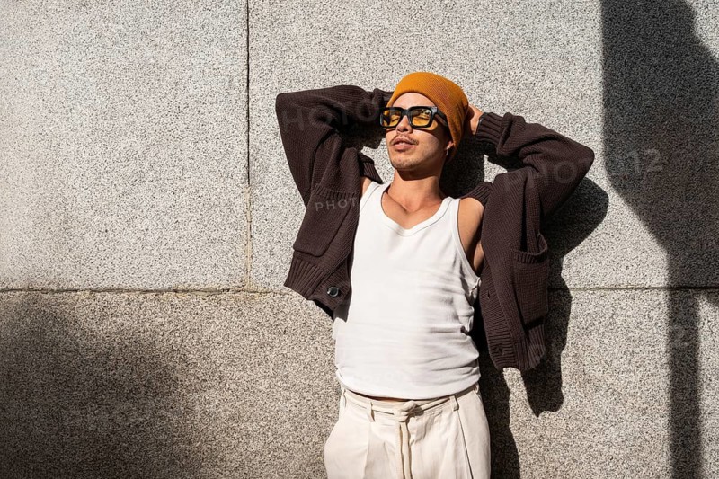 A gay man dressed in casual attire relaxes against a textured wall, basking in sunlight. He wears sunglasses, a mustard beanie, a brown sweater, and white pants, exuding a chilled vibe