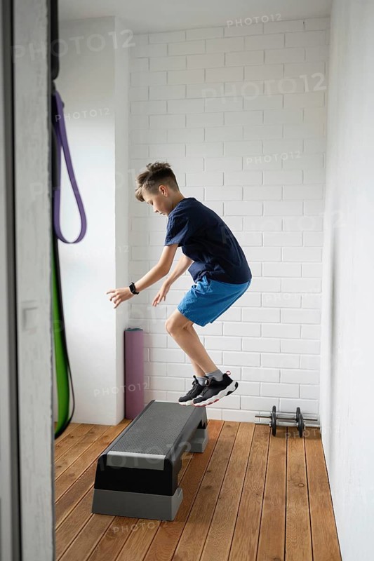 Young boy performing energetic exercises on a step platform indoors. He demonstrates strength and agility in a modern home gym setting, emphasizing active lifestyle commitment