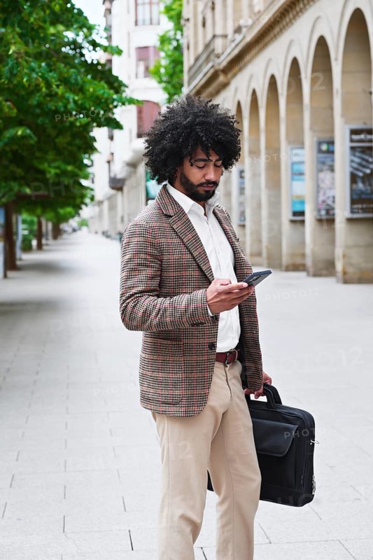 A mixed-race insurance salesman with afro hair checks his smartphone while walking in an urban setting with arched buildings, showcasing a professional and stylish appearance