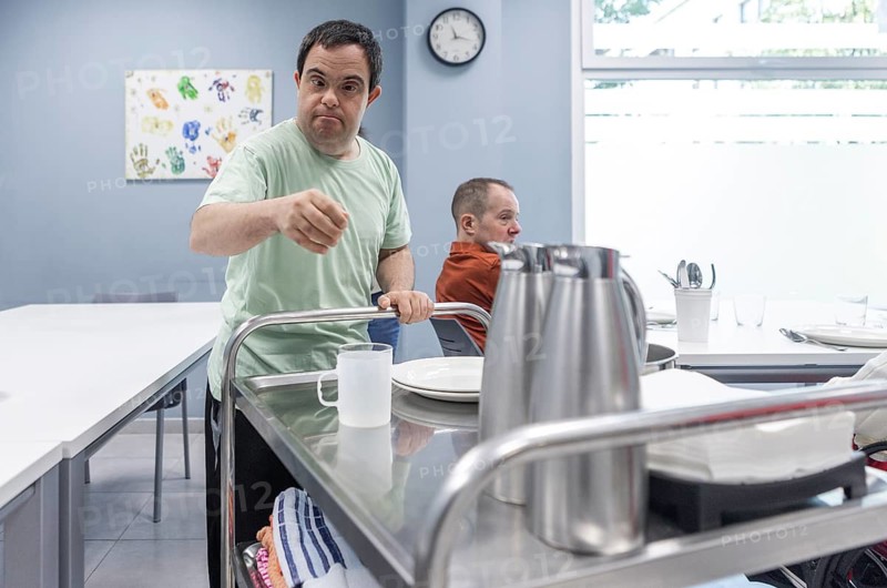 Man with Down syndrome sets the table, placing dishes and cups from a serving cart. The scene highlights independence, inclusion, and the development of life skills in a supportive environment