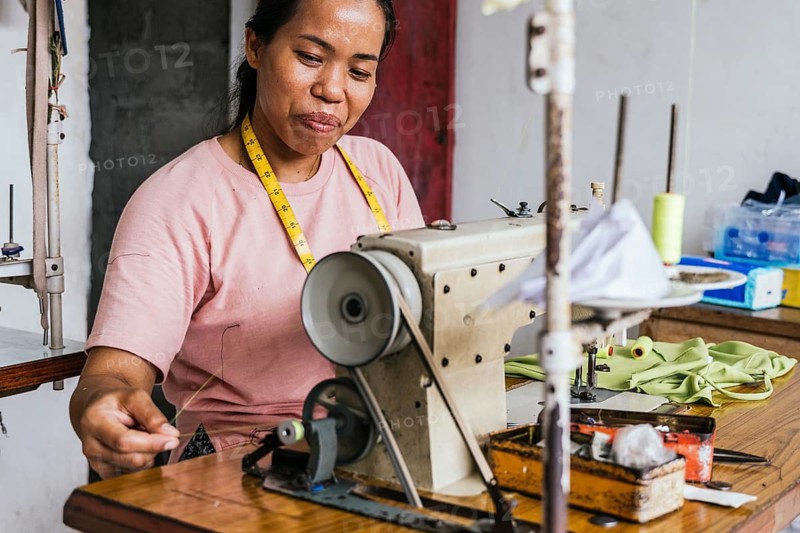 A asian seamstress works diligently at a sewing machine in a tailor shop. Her measuring tape hangs around her neck as she focuses on the task at hand, surrounded by fabrics and tools