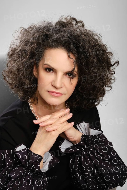 Closeup portrait of senior woman with lush curly hair and black pantsuit posing in studio over grey background