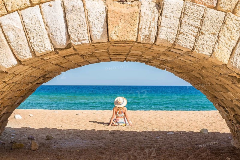 A woman enjoys a peaceful moment on a sandy beach under a stone arch, with the vibrant blue sea stretching beyond. This serene setting captures the beauty of Milos Island