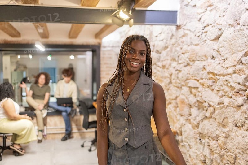 A young professional African woman with braids smiles confidently in a trendy summer coworking space, where colleagues engage in a lively discussion in the background