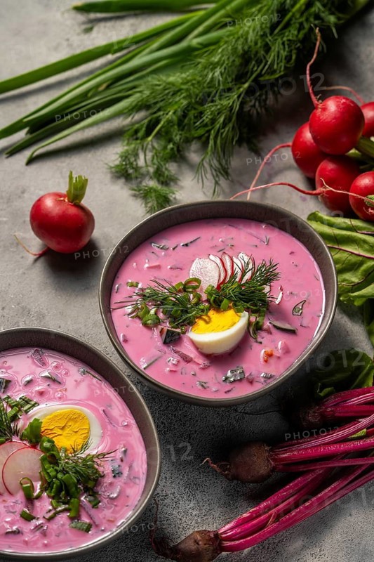 Delicious cold beetroot soup garnished with sliced radishes, hard-boiled eggs, and fresh dill. Surrounded by raw radishes and greens, it offers a refreshing, colorful meal