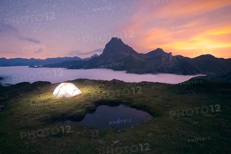A glowing tent sits on a grassy hill under a starlit sky, surrounded by fog covered mountains in the Pyrenees. The serene scene captures the essence of adventure and tranquility in summer