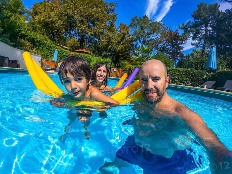Family of three, including a father, mother, and child, smiling at the camera while swimming in a clear blue pool on a sunny day, enjoying summer holidays and refreshing water activities