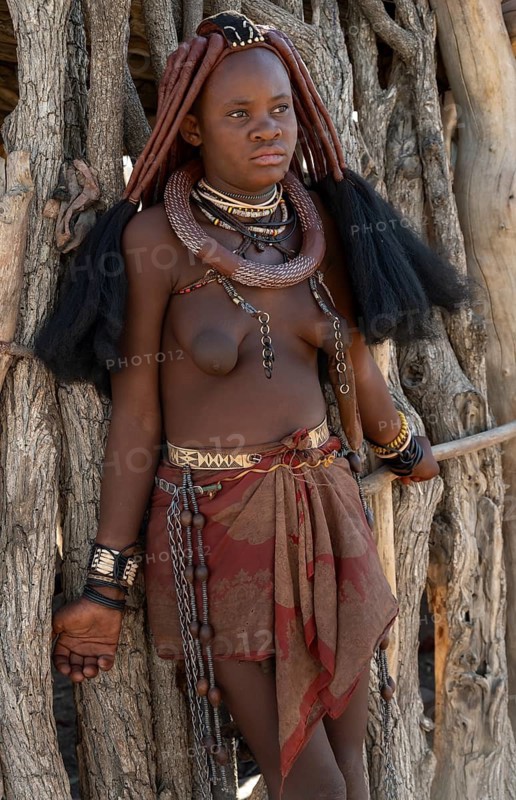Young traditionally dressed Himba woman, leaning against a wooden hut, near Opuwo, Kaokoveld, Kunene, Namibia