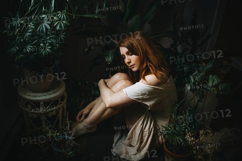 A young woman peacefully sits among lush indoor plants, surrounded by greenery. Her serene expression and relaxed posture evoke a sense of tranquility and introspection