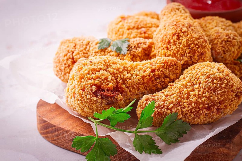 A wooden board holds several golden chicken drumsticks arranged neatly. A small bowl of red dipping sauce is nearby. Fresh parsley adds a touch of color