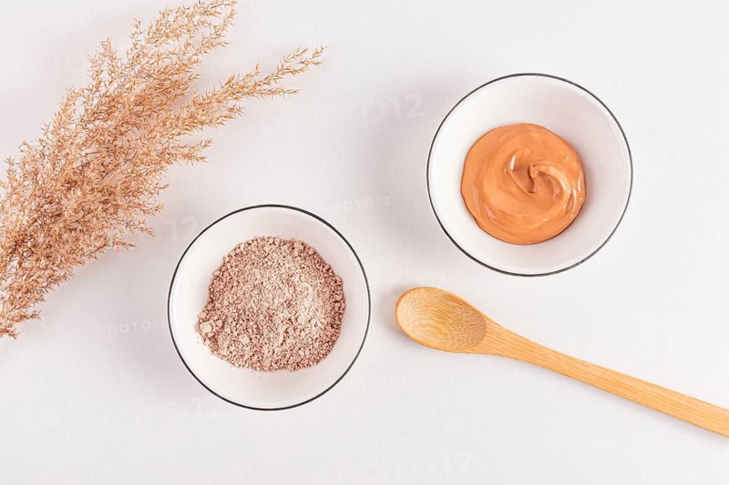 Top view of dry cosmetic clay powder and mixed clay facial mask in white bowls with wooden spoon and dried grass, isolated on a white surface