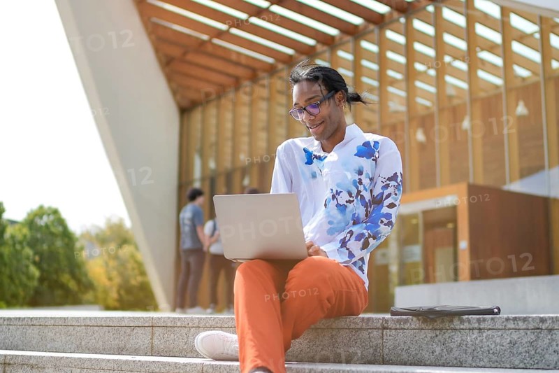 A man in a floral shirt and red pants sits outside, working on a laptop. The scene exudes confidence and pride, set against a modern architectural backdrop