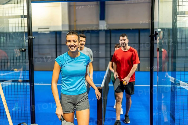 Group of happy young adults, including a smiling woman holding a paddle, entering a vibrant blue indoor court for a competitive padel game, enjoying active lifestyle and teamwork