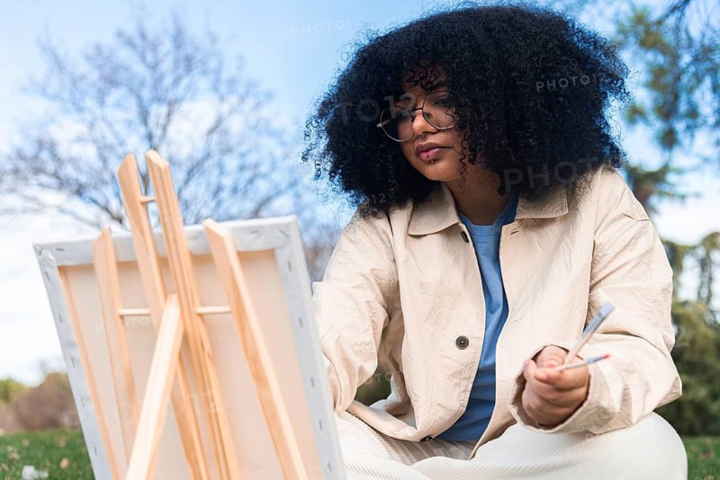 A young black artist with curly hair paints on a canvas in an outdoor setting. Wearing glasses and a beige jacket, they hold brushes, showcasing creativity and focus amid nature