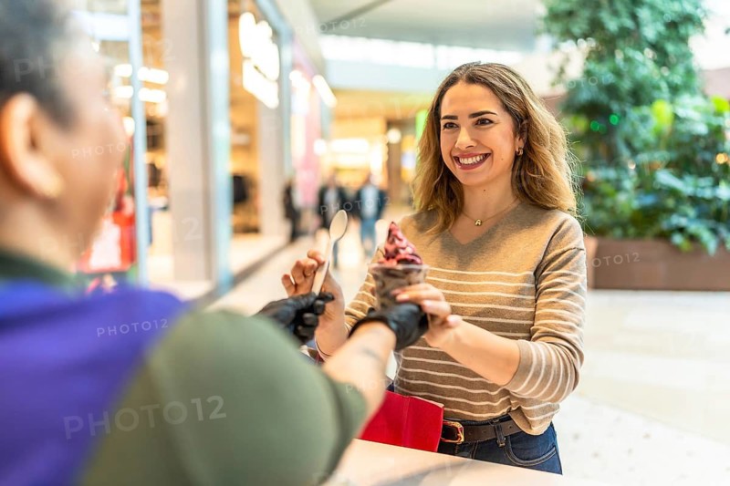 Woman with a beaming smile receiving a cup of delicious ice cream from a gloved vendor, enjoying a sweet treat and excellent customer service during her shopping trip at the modern mall