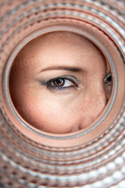 A close-up of a woman's eye peering through the circular opening of a textured pipe, capturing the detail of her makeup, skin texture, and the pipe's intricate pattern