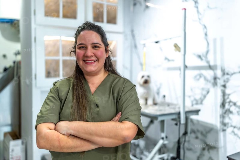 Professional woman vet technician or small business owner standing with arms crossed, smiling at the camera as a dog waits on a grooming table in her pet care salon