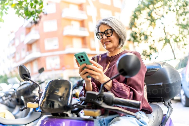 Senior gray haired woman with glasses sitting on a scooter on a sunny city street, smiling as she uses her smartphone for messaging, browsing and navigating apps while commuting