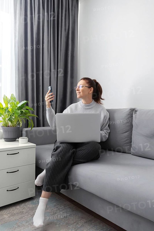 A woman sitting on a comfortable gray sofa, holding a phone and using a laptop for work in a cozy living room. The side table holds a potted plant and a cup, enhancing the serene atmosphere