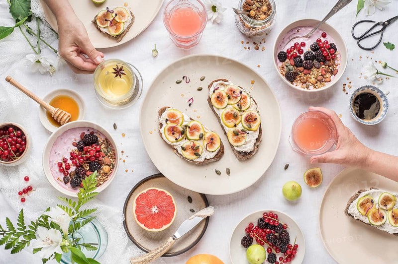 From above anonymous people taking cups of fresh healthy beverages from table with assorted food for breakfast