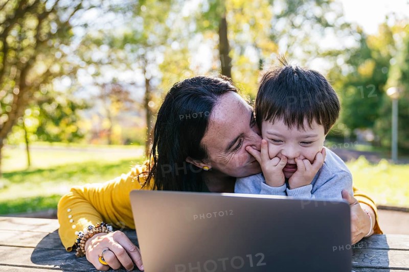 A loving mother and her child with Down syndrome enjoy quality time outdoors, playing and learning with a laptop. Sunlight filters through trees, creating a warm, joyful scene